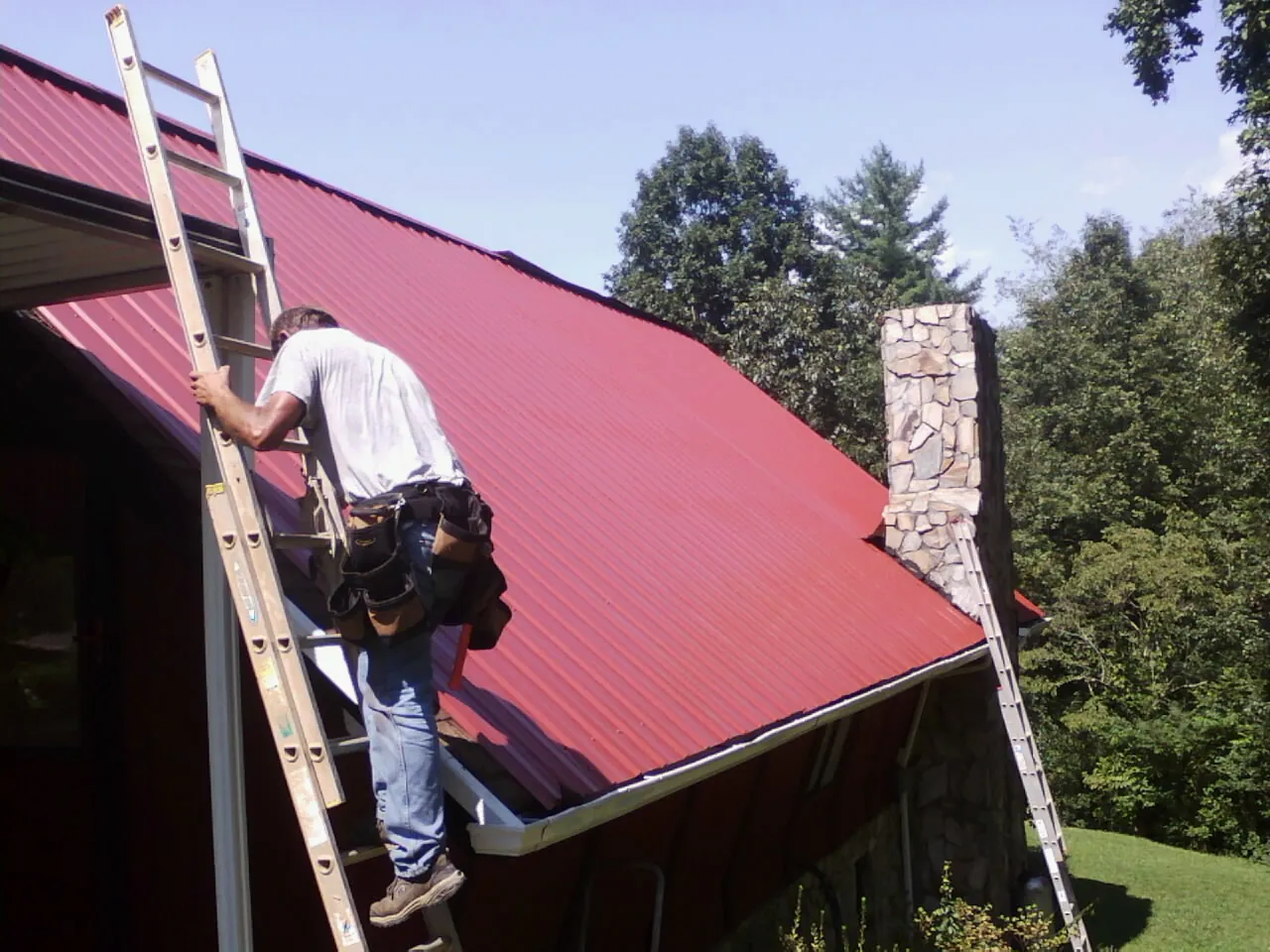 Roofer on a ladder beside a red metal roof and stone chimney — Soffit & Fascia Repair by Roofing Brothers in Noxen